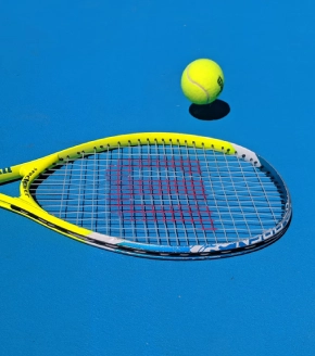 The photo shows a close-up of a tennis racket and ball on a bright blue tennis court, suggesting the presence of sports facilities at Dosti West County Thane Balkum